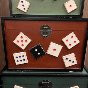 Puede incluir: Tres cajas decorativas de madera, apiladas, con una variedad de dados esparcidos encima. Las cajas son marrones y verdes con ribetes negros. Los dados son negros y blancos con puntos rojos y azules.