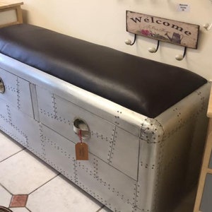 Puede incluir: Un banco de almacenamiento de metal plateado con un asiento superior de cuero marrón. El banco tiene dos cajones con remaches y una manija de metal. El banco está en una habitación con pisos de baldosas.