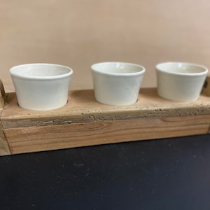 Puede incluir: Una jardinera de madera con tres macetas de cerámica blanca. La jardinera está hecha de madera natural con un acabado rústico.