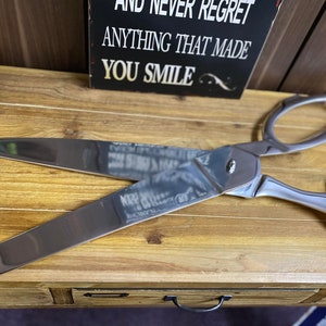 May include: Large, shiny silver scissors with loop handles. The blades are open, revealing a sharp edge. A small potted plant and a sign with the words "AND NEVER REGRET ANYTHING THAT MADE YOU SMILE" are in the background. The scissors are on a wooden surface.