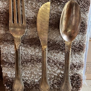 Pode incluir: Conjunto de três talheres decorativos de metal grandes: um garfo, uma faca e uma colher. O garfo tem quatro dentes, a faca tem uma lâmina curva e a colher tem uma tigela arredondada. Acabamento dourado antigo e detalhes ornamentais.
