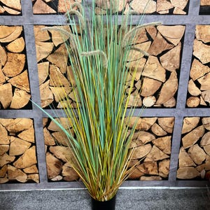 Puede incluir: Una planta alta de hierba ornamental artificial en una maceta negra. Las hojas son una mezcla de verde, amarillo y marrón, con penachos plumosos. El fondo presenta una pared de troncos de madera apilados.