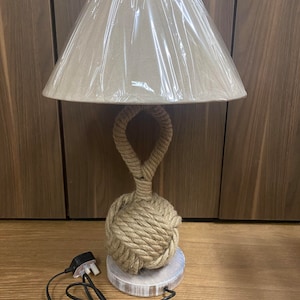 Puede incluir: Una pantalla de lámpara beige con una base de cuerda en forma de nudo. La lámpara está sobre una base de madera y tiene un cable negro.