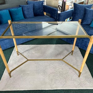 May include: A rectangular coffee table with a clear glass top and a gold-coloured metal frame. The legs are light-coloured wood with gold-coloured metal accents. The table is set on a light-coloured rug.