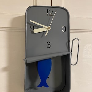 May include: A grey wall clock in the shape of a sardine tin, featuring a blue fish design. The clock has white hands and the numbers 3, 6, 9, and 12 are visible. It's mounted on a white door.