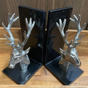 May include: A pair of black bookends featuring silver deer head sculptures. The deer heads have detailed antlers and a realistic appearance.