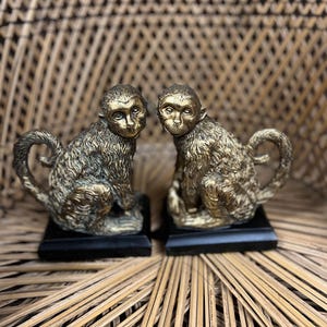 Könnte beinhalten: Ein Paar bronzefarbene Affenfiguren, jeweils auf einem schwarzen rechteckigen Sockel sitzend. Die Affen haben eine detaillierte Fellstruktur und sind mit eingerollten Schwänzen posiert. Der Hintergrund ist ein geflochtenes Korbgeflechtmuster.
