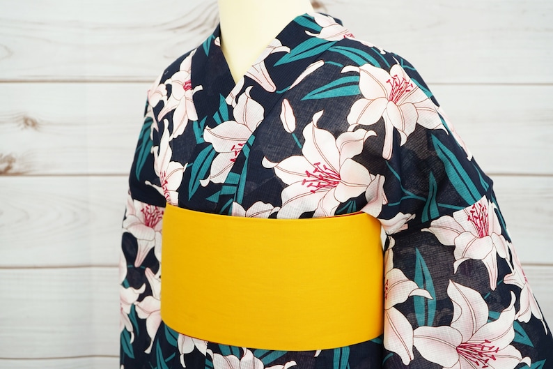 Puede incluir: Un yukata azul marino con un estampado floral de lirios blancos y una faja amarilla. El yukata es una prenda tradicional japonesa que se usa durante los meses de verano.