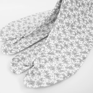 Può includere: Un paio di calzini tabi bianchi con un delicato motivo floreale nero. I calzini sono progettati con una punta divisa, un design tradizionale giapponese. Il tessuto è morbido e sembra essere cotone o un materiale simile.