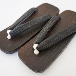 May include: A pair of traditional Japanese geta sandals. The wooden platforms are dark brown with black fabric straps. The straps are attached to the platform with white cords. The sandals are displayed on a white background.