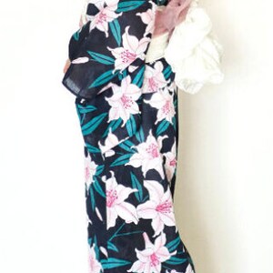 Puede incluir: Una mujer vistiendo una yukata negra y verde con un estampado floral de lirios blancos. La yukata tiene un cintur&oacute;n obi rosa y blanco. La mujer lleva sandalias de madera.