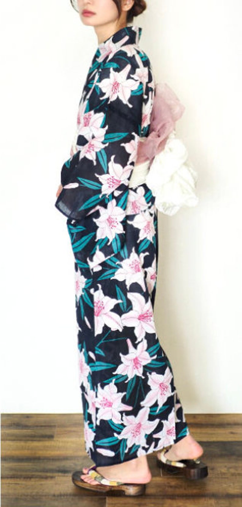 Puede incluir: Una mujer vistiendo una yukata negra y verde con un estampado floral de lirios blancos. La yukata tiene un cintur&oacute;n obi rosa y blanco. La mujer lleva sandalias de madera.