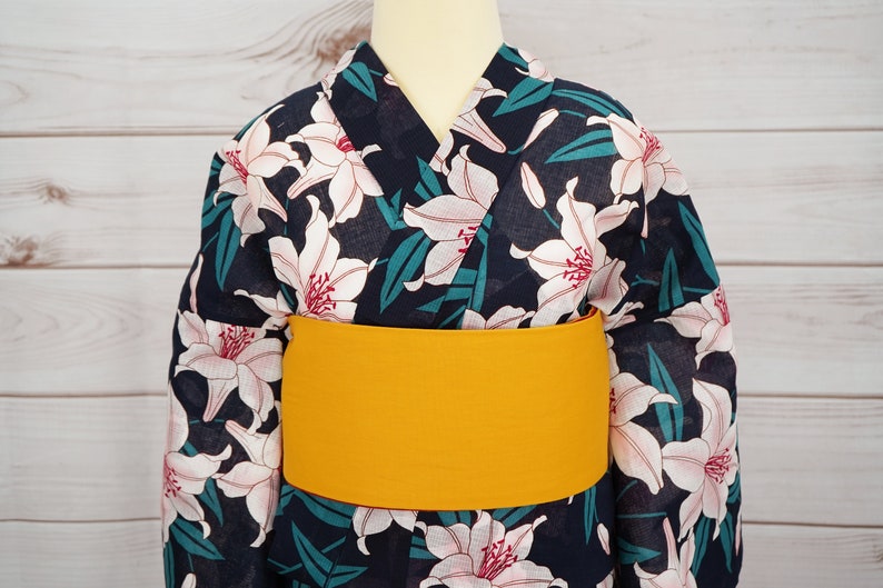 Puede incluir: Un yukata azul marino con un estampado floral de lirios blancos y una faja obi amarilla.