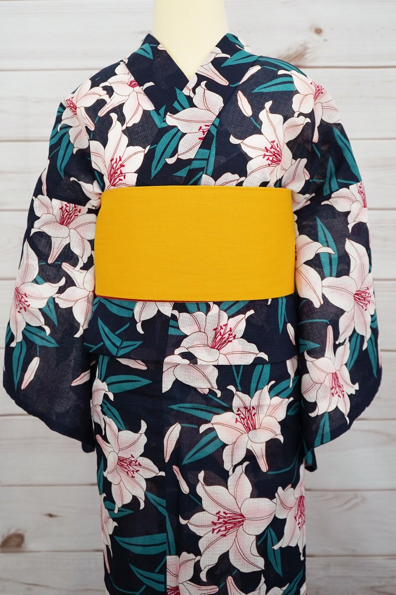 Puede incluir: Un yukata azul marino con un estampado floral de lirios blancos y una faja amarilla. El yukata es una prenda tradicional japonesa que se usa durante los meses de verano.