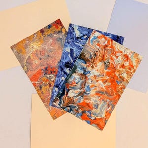 May include: Three abstract art prints with vibrant colors. One print features orange, red, and white swirls. Another showcases blue and white patterns, and the third combines orange, brown, and white. The prints are arranged on a pastel background.