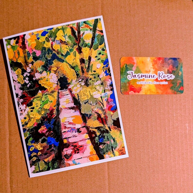 May include: A rectangular art print featuring an impressionistic painting of a path surrounded by vibrant yellow, green, pink, and blue foliage. Beside it is a small rectangular business card with the text Jasmine Rose multimedia fine artist.