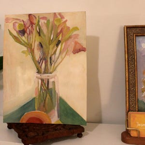 May include: An original painting of a bouquet of flowers in a clear glass jar. The flowers are pink and yellow, with green stems and leaves. The painting is on a small canvas, displayed on a wooden stand.