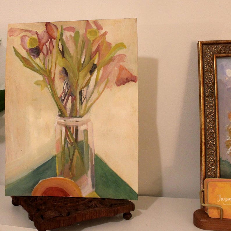 May include: An original painting of a bouquet of flowers in a clear glass jar. The flowers are pink and yellow, with green stems and leaves. The painting is on a small canvas, displayed on a wooden stand.
