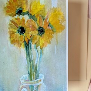 Puede incluir: Pintura a la acuarela de girasoles en un jarr&oacute;n de cristal. Los girasoles son amarillos con centros oscuros y tallos verdes. El jarr&oacute;n es transparente y el fondo es de un color suave y neutro. Un sobre beige est&aacute; al lado de la pintura.