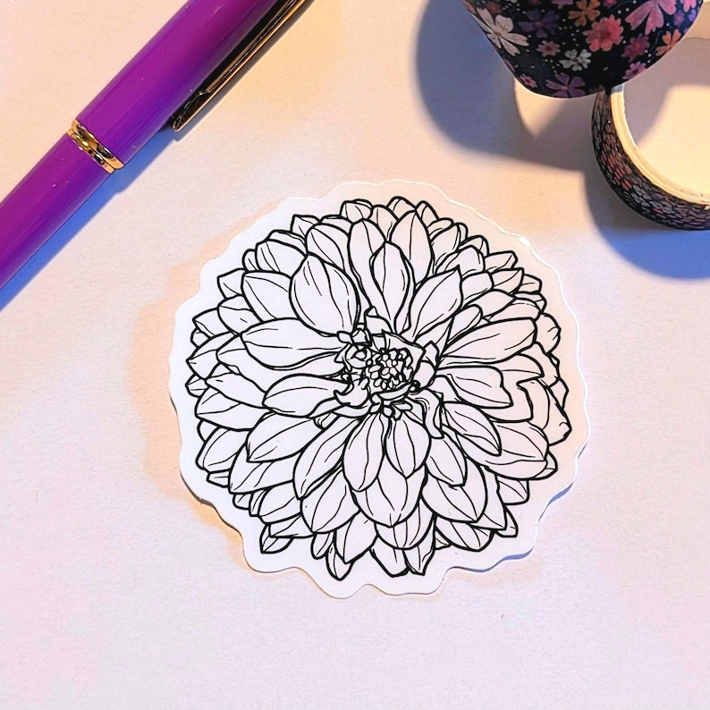 Puede incluir: Una pegatina floral en blanco y negro con un contorno detallado de una flor de dalia. La pegatina tiene un borde blanco y est&aacute; colocada sobre una superficie clara. Tambi&eacute;n se ven un bol&iacute;grafo morado y cinta adhesiva con motivos florales.
