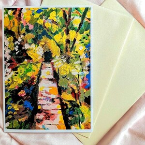 May include: A rectangular greeting card featuring an impressionistic painting of a stone pathway surrounded by vibrant yellow, green, and blue foliage. The card is accompanied by a plain, light-colored envelope, both displayed on a soft fabric surface.