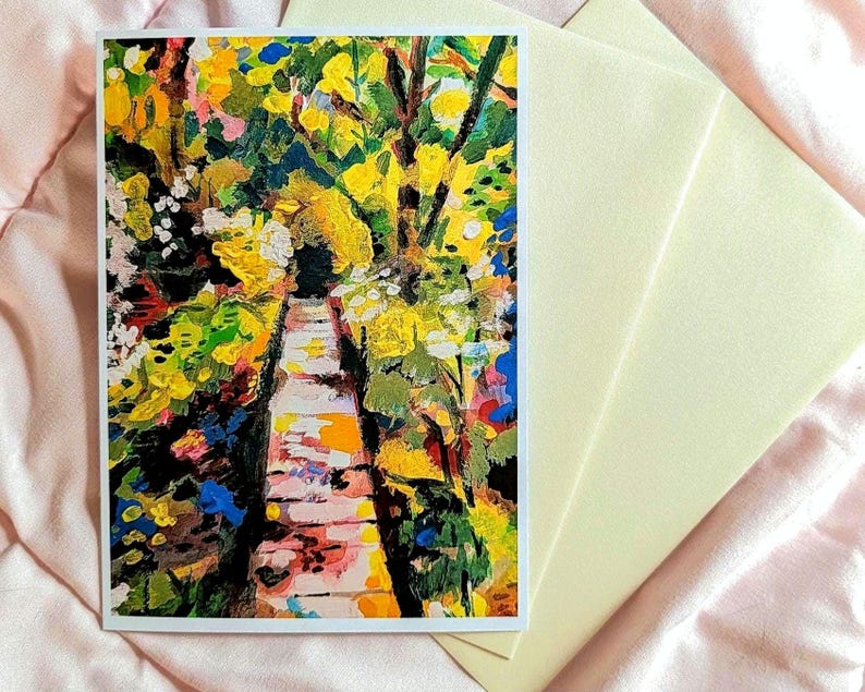 May include: A rectangular greeting card featuring an impressionistic painting of a stone pathway surrounded by vibrant yellow, green, and blue foliage. The card is accompanied by a plain, light-colored envelope, both displayed on a soft fabric surface.
