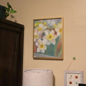 Puede incluir: Una pintura enmarcada de narcisos blancos y amarillos con tallos y hojas verdes. La obra de arte est&aacute; en un marco dorado y cuelga de una pared beige. La pintura est&aacute; iluminada por una l&aacute;mpara cercana.