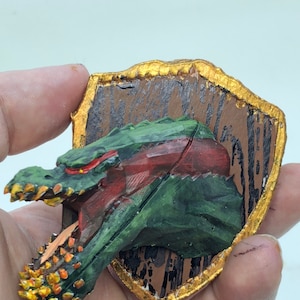 May include: A hand-painted dragon head sculpture mounted on a wooden plaque. The dragon features green scales, red accents, and a golden-yellow mouth. The plaque has a brown background with a gold-colored border.