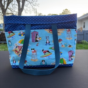 May include: A blue tote bag with a beach-themed pattern featuring dogs surfing, sunbathing, and playing. The bag has a navy blue polka dot top and straps, perfect for carrying beach essentials. The bag measures approximately 16 inches wide and 12 inches tall.