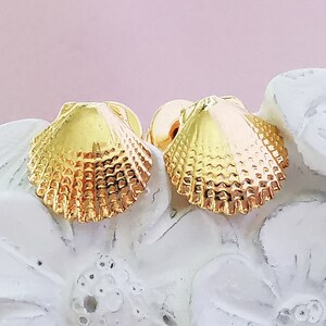 Puede incluir: Un par de pendientes de color dorado en forma de concha. Los pendientes son pequeños y delicados, y tienen una superficie texturizada.