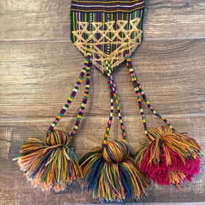 May include: A woven textile strap featuring a geometric pattern in green, blue, yellow, and pink, ending in a pointed section with gold-coloured embroidery. Three braided cords extend from the point, each finished with a large, multi-coloured yarn tassel.