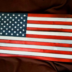 May include: A wooden American flag with red, white, and blue paint. The flag is made of individual wooden planks, creating a rustic look. The stars are painted in white on a blue background.