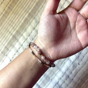 May include: Two bracelets on a wrist. One bracelet is made of small pink, white, and gold beads. The other bracelet is made of larger pink and brown beads.