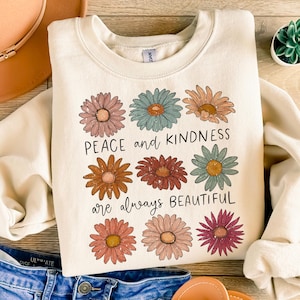 K&ouml;nnte beinhalten: Ein cremefarbener Pullover mit einem grafischen Design aus bunten Blumen und dem Text "Peace and kindness are always beautiful".