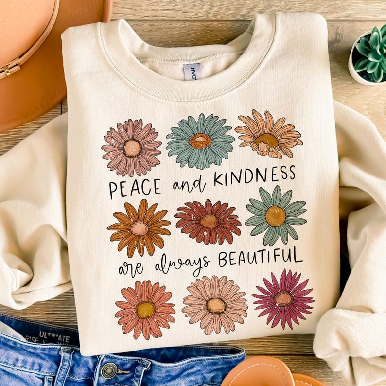 K&ouml;nnte beinhalten: Ein cremefarbener Pullover mit einem grafischen Design aus bunten Blumen und dem Text "Peace and kindness are always beautiful".