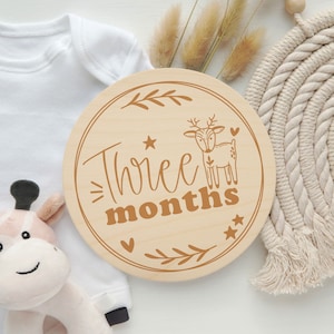 May include: A round wooden sign with a brown deer and the text "Three months" in brown lettering. The sign is decorated with small hearts and stars.
