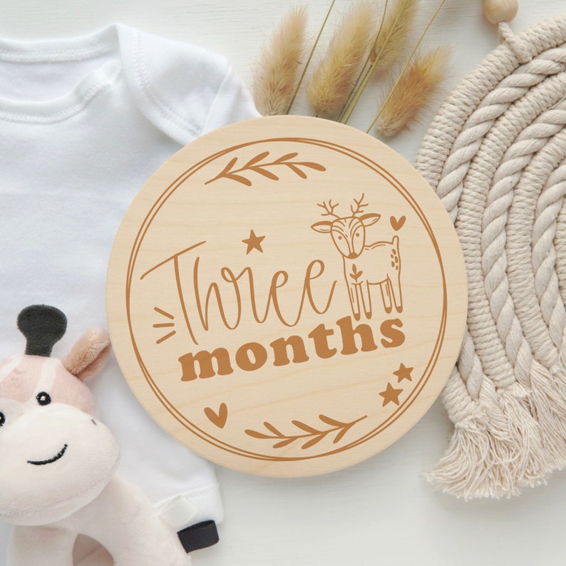 May include: A round wooden sign with a brown deer and the text "Three months" in brown lettering. The sign is decorated with small hearts and stars.