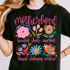 Op de afbeelding: Zwart t-shirt met het woord "Motherhood" in roze script, omringd door kleurrijke bloemenontwerpen. Daaronder staan de woorden "beautiful chaotic important blessed challenging worth it" in roze script.