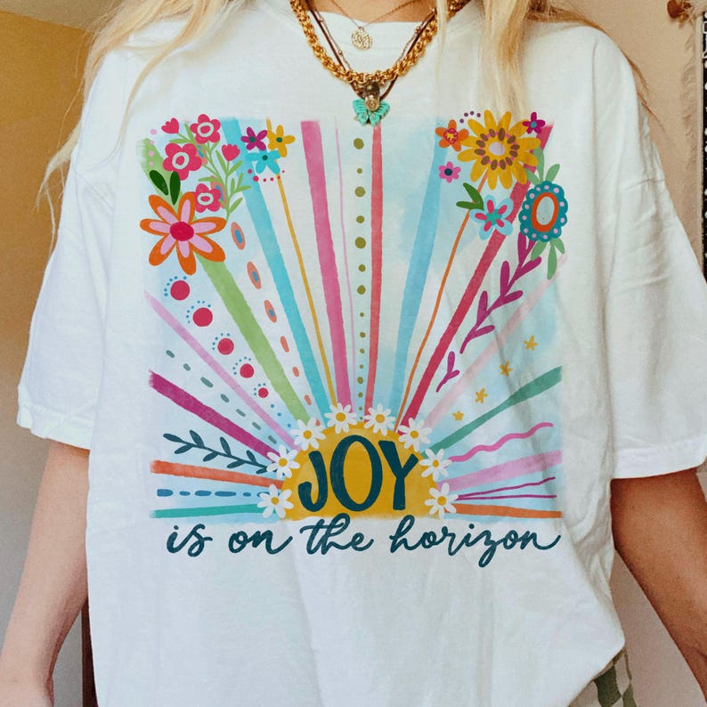 May include: White t-shirt with a colorful graphic design of a sun with rays and flowers. The text "JOY is on the horizon" is printed below the sun.