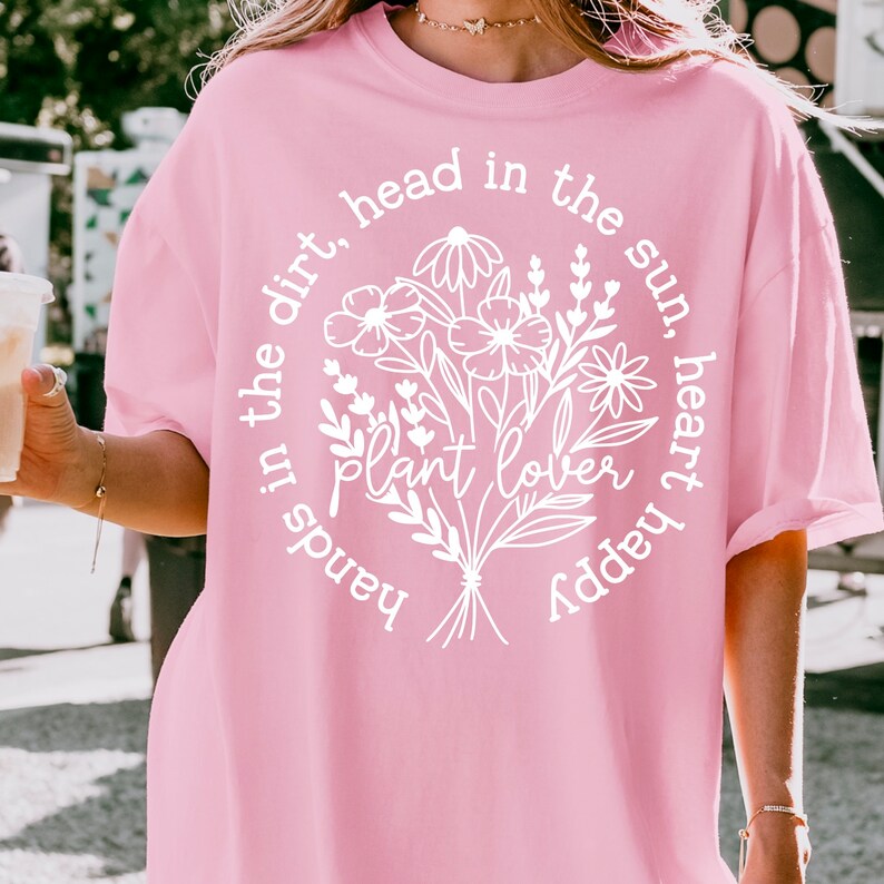 May include: Pink oversized t-shirt with a white graphic design of flowers and the text "Hands in the dirt, head in the sun, heart happy plant lover".