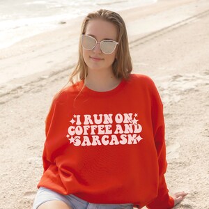 May include: A woman wearing a red sweatshirt with the text "I run on coffee and sarcasm" in white retro font. She is wearing sunglasses and is standing on a sandy beach.