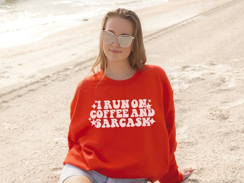 May include: A woman wearing a red sweatshirt with the text "I run on coffee and sarcasm" in white retro font. She is wearing sunglasses and is standing on a sandy beach.