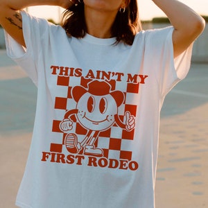 May include: White t-shirt with a red graphic of a cartoon character wearing a cowboy hat and the text "This ain't my first rodeo".