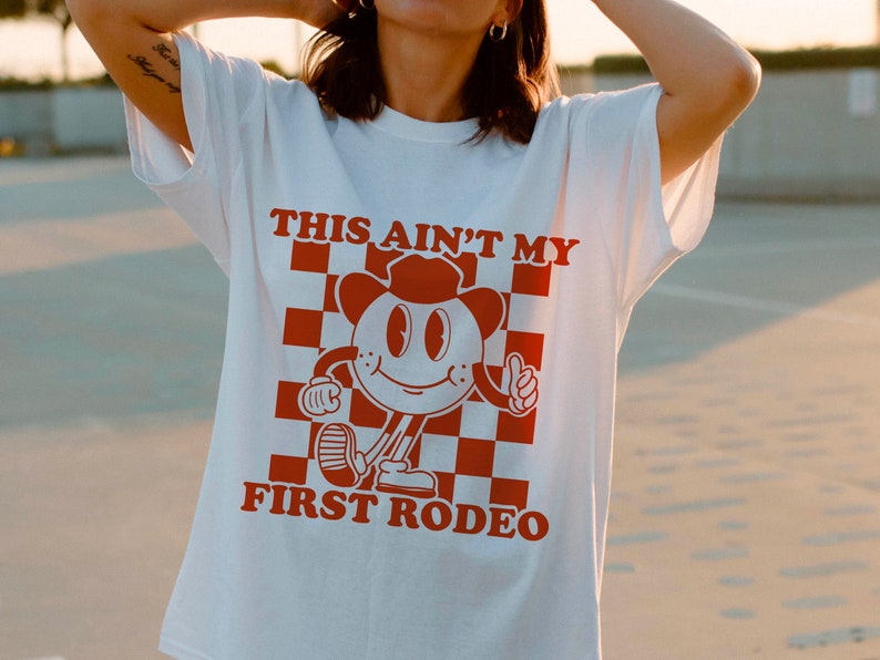 May include: White t-shirt with a red graphic of a cartoon character wearing a cowboy hat and the text "This ain't my first rodeo".