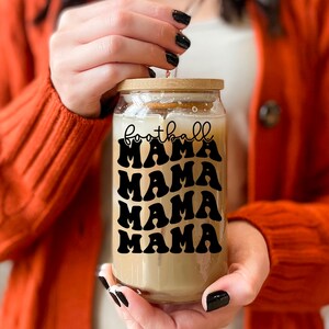May include: A clear glass jar with a wooden lid and a black and white graphic that says "football mama" repeated four times. The jar is filled with a light brown liquid.