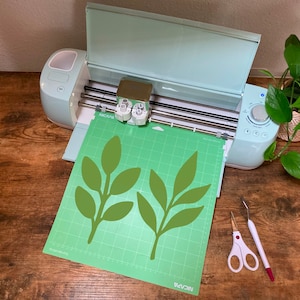May include: A green cutting mat with two green leaf designs on it, placed in a light blue Cricut Explore Air 2 cutting machine. The machine is open and the cutting blade is visible. A pair of silver scissors and a white and red tool are on the wooden table next to the machine.