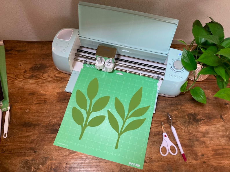 May include: A green cutting mat with two green leaf designs on it, placed in a light blue Cricut Explore Air 2 cutting machine. The machine is open and the cutting blade is visible. A pair of silver scissors and a white and red tool are on the wooden table next to the machine.
