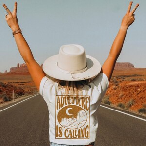 May include: A person wearing a white t-shirt with a brown graphic that says "Adventure is Calling" and a white hat with a brown band. The person is standing on a road with red dirt on either side.