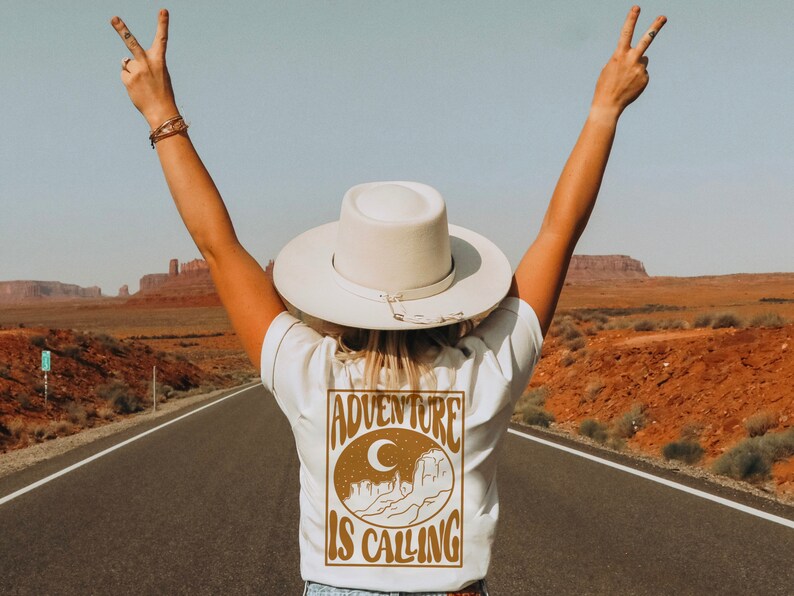 May include: A person wearing a white t-shirt with a brown graphic that says "Adventure is Calling" and a white hat with a brown band. The person is standing on a road with red dirt on either side.