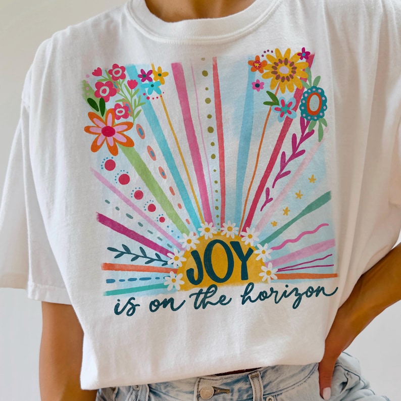 May include: White t-shirt with a colorful graphic design of a sun with the text "JOY is on the horizon".
